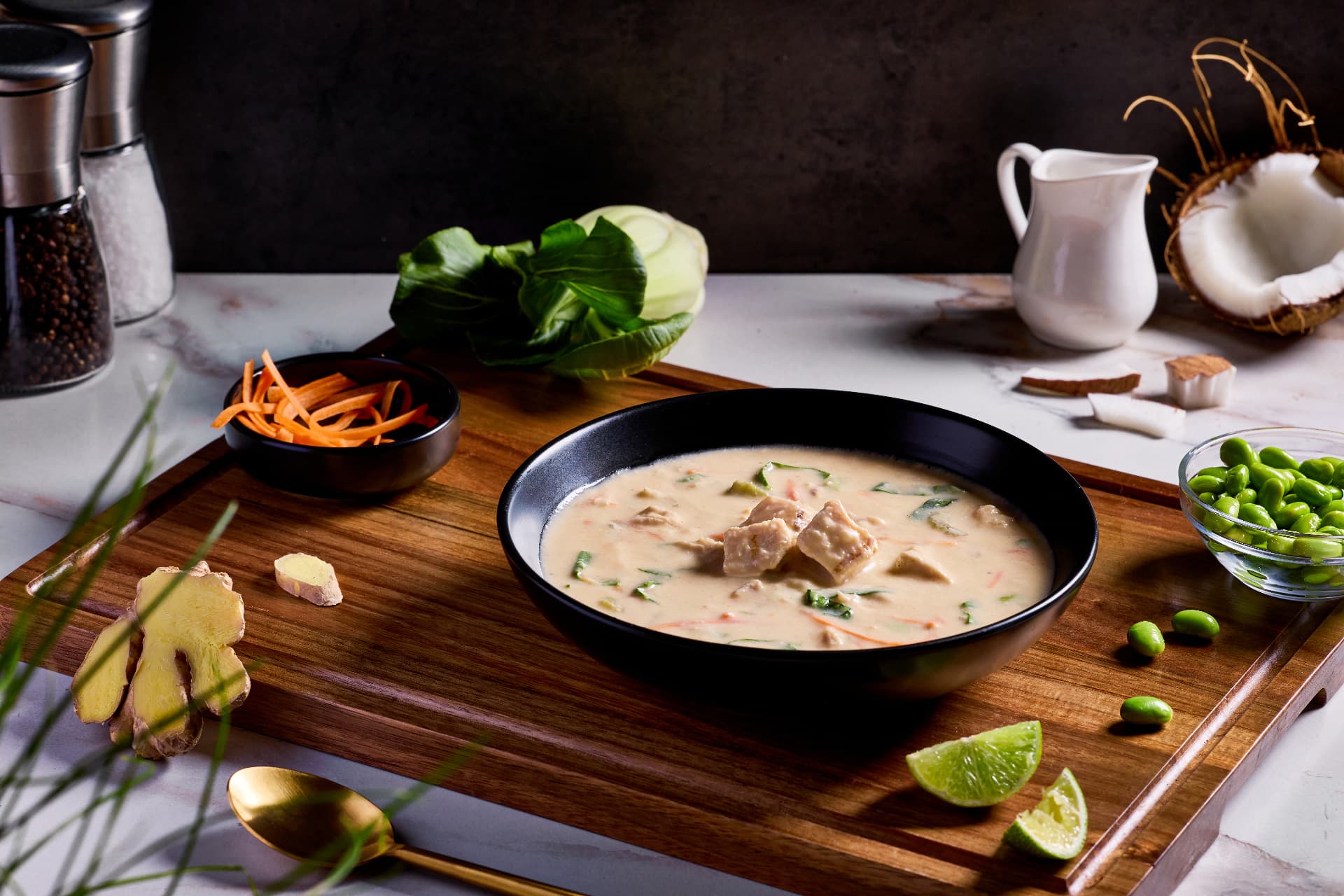 A bowl of coconut chicken soup with fresh ginger, lime, bok choy and edamame on a wooden serving board