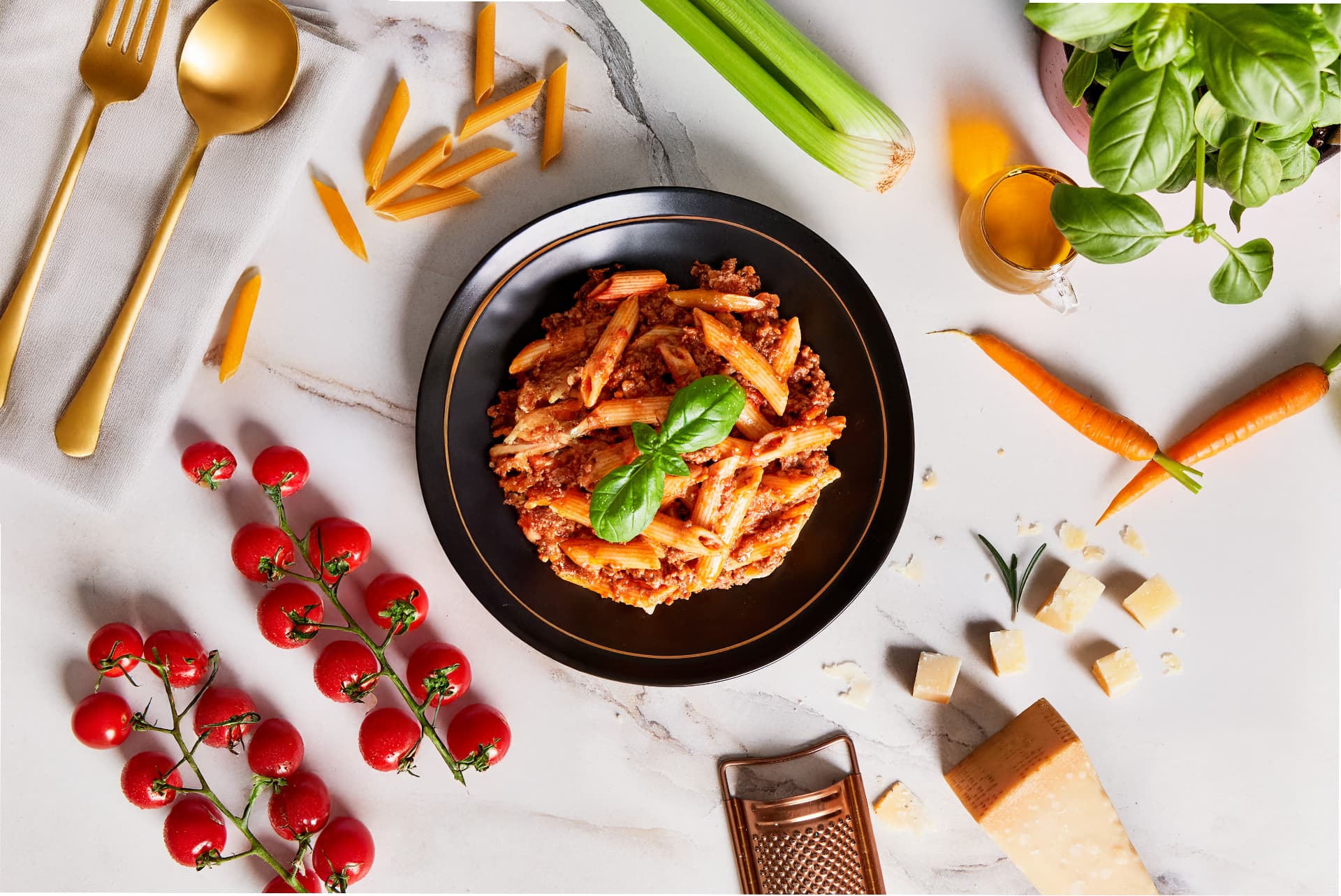Overhead view of penne bolognese plated on a black dish surrounded by tomatoes, basil, carrots and parmesan