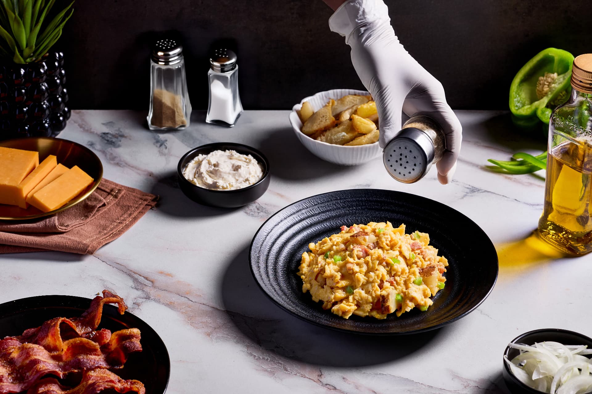 A plated Denver-style scrambled egg dish being finished with seasoning beside bacon, cheddar and peppers