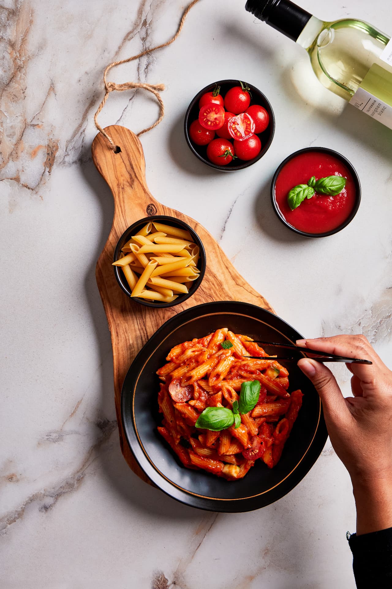 A hand plating penne arrabbiata with tongs beside dry penne, cherry tomatoes and tomato sauce